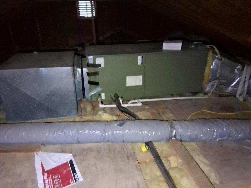 Completed AC system estimate in attic space. Documented existing green air handler unit positioned on wooden platform with exposed ductwork and insulation. Photographed equipment dimensions, duct configurations, and current installation conditions including flexible ductwork and fiberglass insulation placement for estimate preparation.