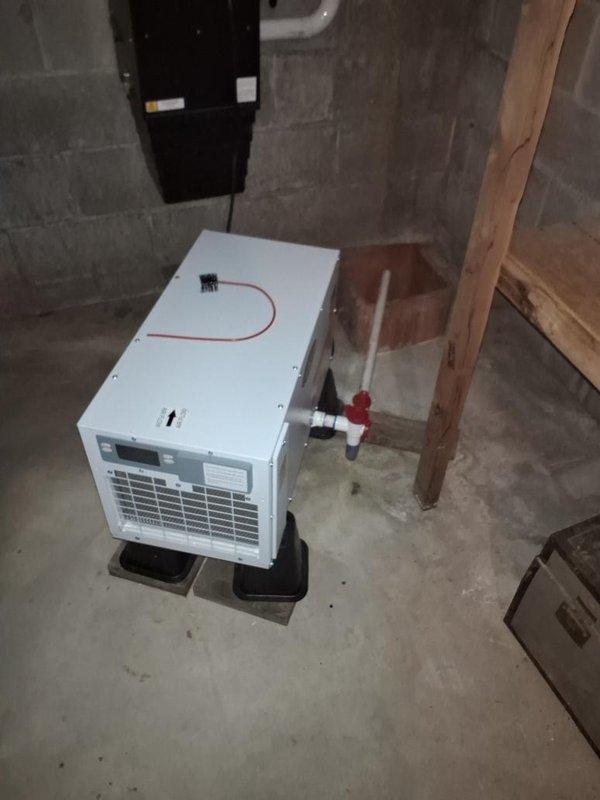 Responded to service call for leaking dehumidifier in basement utility area. Upon arrival, inspected white portable dehumidifier unit positioned on black plastic supports in unfinished mechanical room with concrete block walls and floor. Examined unit for source of water leak, checking drain connections, condensate pan, and overflow mechanisms. Verified proper drainage routing through connected PVC piping system and confirmed unit was level on supporting platform to ensure proper condensate flow. Tested unit operation and monitored for leakage during run cycle to verify repair completion.