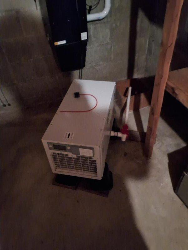 Responded to service call for leaking dehumidifier in basement utility area. Upon arrival, inspected white portable dehumidifier unit positioned on black plastic supports in unfinished mechanical room with concrete block walls and floor. Examined unit for source of water leak, checking drain connections, condensate pan, and overflow mechanisms. Verified proper drainage routing through connected PVC piping system and confirmed unit was level on supporting platform to ensure proper condensate flow. Tested unit operation and monitored for leakage during run cycle to verify repair completion.