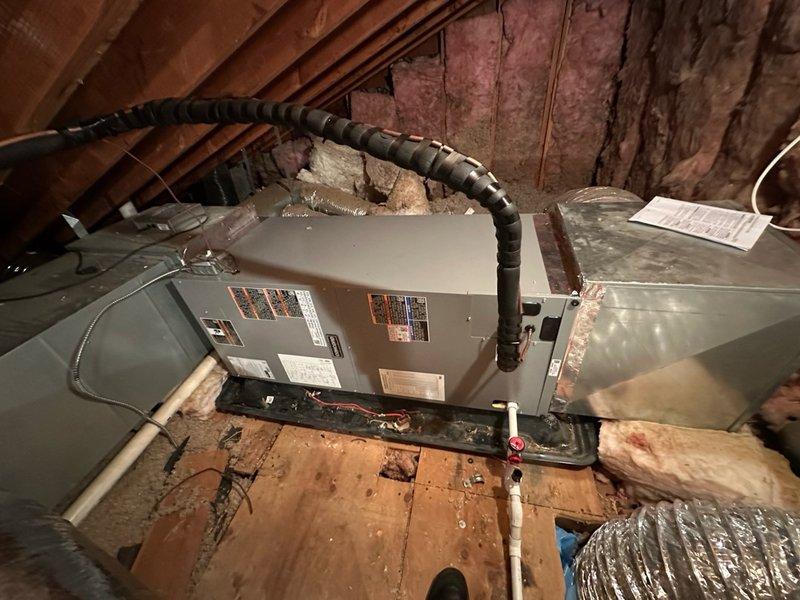 Completed installation of Ultimate Comfort heat pump system in attic space, providing high-efficiency heating and cooling with dual fuel capability when integrated with existing gas or oil heating system. Installed gray metal air handler unit horizontally between exposed wooden joists with proper mounting and secure placement. Connected black flexible ductwork to air handler with additional gray metal ductwork components installed on both sides of unit for complete air distribution throughout the space. Routed white electrical conduit along wooden joists with secure clips for proper power supply to equipment. Verified all manufacturer specifications and technical requirements per unit labels and diagrams. Installation includes insulated flex ducts with proper sealing, cylindrical humidifier component for enhanced comfort control, and all necessary piping, valves, and connecting hardware with red-handled fittings for system operation. System positioned to maximize efficiency while worki