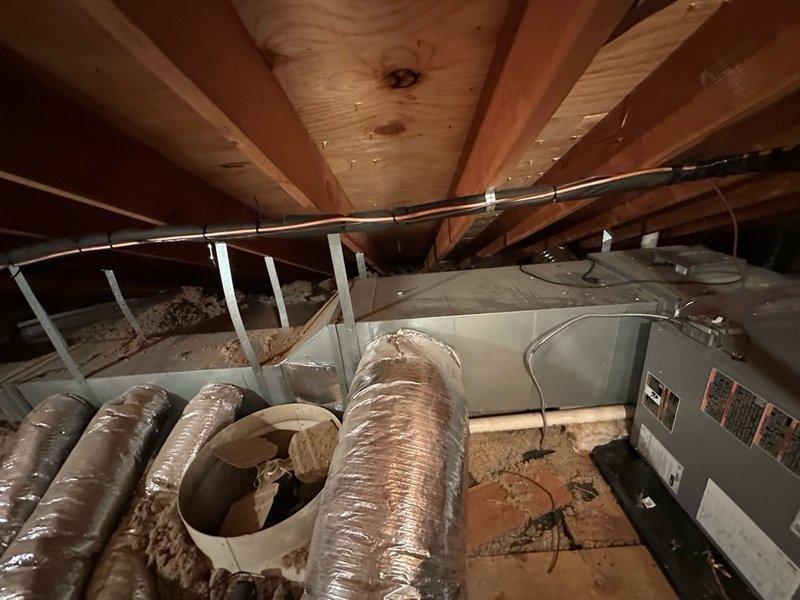 Completed installation of Ultimate Comfort heat pump system in attic space, providing high-efficiency heating and cooling with dual fuel capability when integrated with existing gas or oil heating system. Installed gray metal air handler unit horizontally between exposed wooden joists with proper mounting and secure placement. Connected black flexible ductwork to air handler with additional gray metal ductwork components installed on both sides of unit for complete air distribution throughout the space. Routed white electrical conduit along wooden joists with secure clips for proper power supply to equipment. Verified all manufacturer specifications and technical requirements per unit labels and diagrams. Installation includes insulated flex ducts with proper sealing, cylindrical humidifier component for enhanced comfort control, and all necessary piping, valves, and connecting hardware with red-handled fittings for system operation. System positioned to maximize efficiency while worki