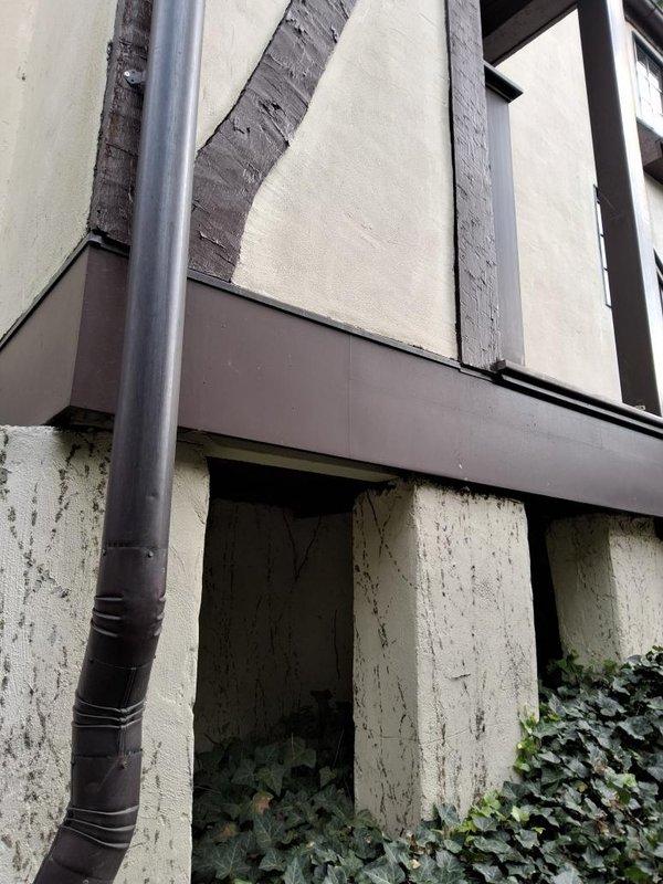 Completed estimate for make-up air system installation. Assessed exterior building conditions including Tudor-style architecture with stucco panels, foundation wall with weathering and vegetation growth, and existing drainage system. Documented current state of building envelope, identified areas of water damage and staining on lower wall surfaces, and evaluated ventilation requirements. Photographed site conditions to support equipment sizing and installation planning for proposed make-up air unit.