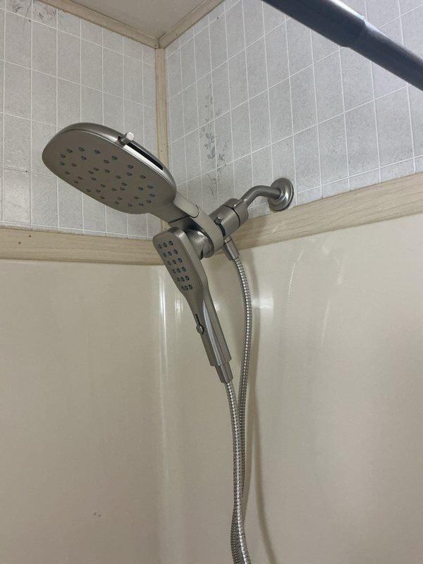 Completed installation of a dual-head shower system in bathroom with main fixed showerhead and secondary handheld unit, both in brushed nickel finish. Accessed and worked on plumbing connections within wall cavity, properly connecting the brass mixing valve to hot (red) and cold (blue) water supply lines. Documented installation with photos showing both the completed fixture and the properly connected plumbing components behind the wall.