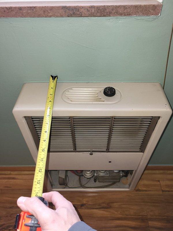 Performed inspection and evaluation of a non-functional 67-year-old wall-mounted gas space heater in the back bedroom. Unit is approximately 15-16 inches wide by 13-14 inches high, mounted on a mint green wall above wooden flooring. Measurements were taken and documentation photos captured to prepare replacement estimate for a new gas-powered wall heater with exterior venting capabilities.