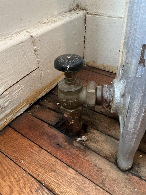 Inspected basement boiler system and identified multiple radiator leaks. Found significant water damage to wooden flooring and wall discoloration beneath affected radiator valves. Observed corrosion and deterioration on brass valves and connecting pipes, which were the source of the leakage.