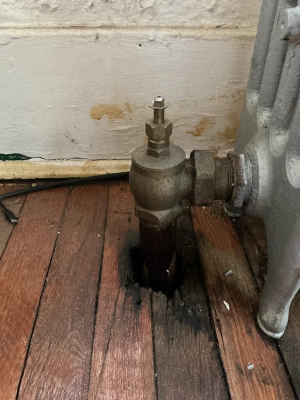 Inspected basement boiler system and identified multiple radiator leaks. Found significant water damage to wooden flooring and wall discoloration beneath affected radiator valves. Observed corrosion and deterioration on brass valves and connecting pipes, which were the source of the leakage.