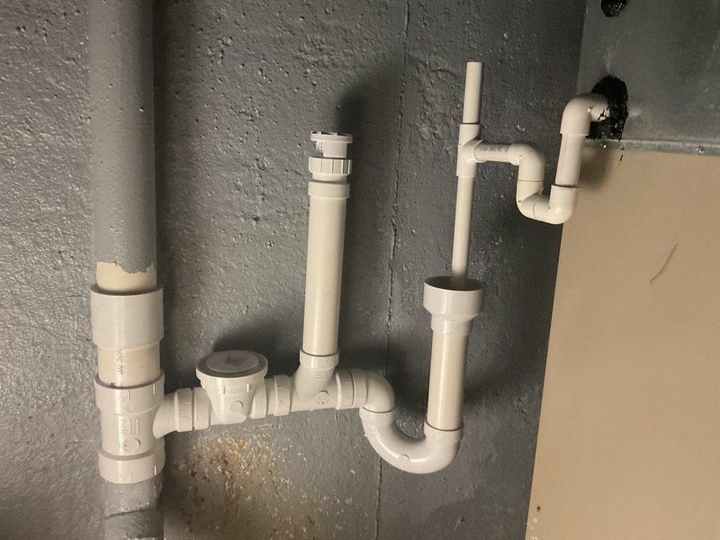 Installed new PVC drainage system on concrete wall, including P-trap configuration, vertical drain pipes with air admittance valve, and proper elbow joints. Sealed pipe wall penetration with black insulation material and confirmed proper drainage functionality after installation.