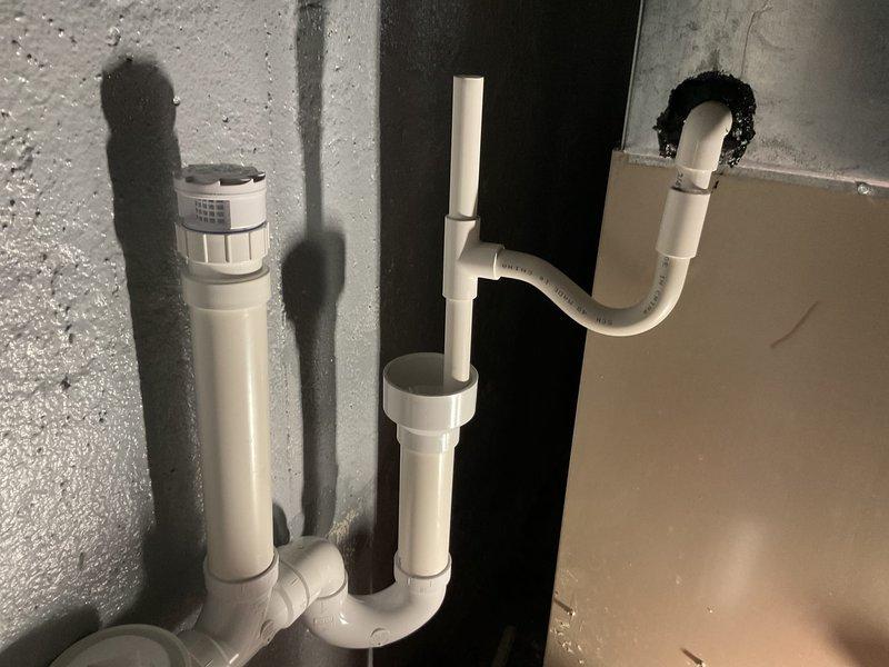 Installed new PVC drainage system on concrete wall, including P-trap configuration, vertical drain pipes with air admittance valve, and proper elbow joints. Sealed pipe wall penetration with black insulation material and confirmed proper drainage functionality after installation.