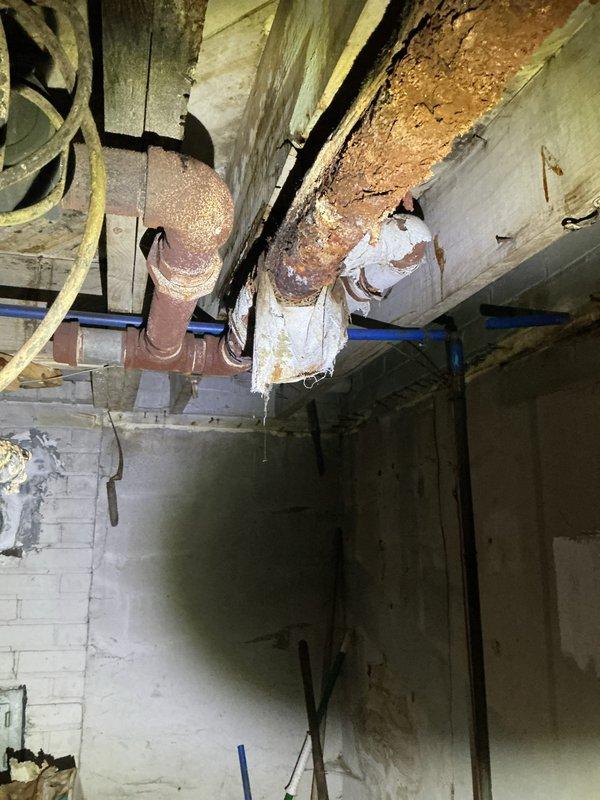 Responded to homeowner's report of burst pipe connected to hot water boiler. Inspection revealed severe corrosion and deterioration of ceiling pipes in basement, with significant water damage evident from leaking. Primary issue identified was a completely failed pipe section with advanced rust damage, causing water to leak onto surrounding structure and creating extensive staining on ceiling and walls.