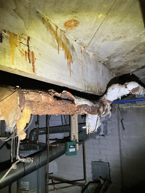 Responded to homeowner's report of burst pipe connected to hot water boiler. Inspection revealed severe corrosion and deterioration of ceiling pipes in basement, with significant water damage evident from leaking. Primary issue identified was a completely failed pipe section with advanced rust damage, causing water to leak onto surrounding structure and creating extensive staining on ceiling and walls.