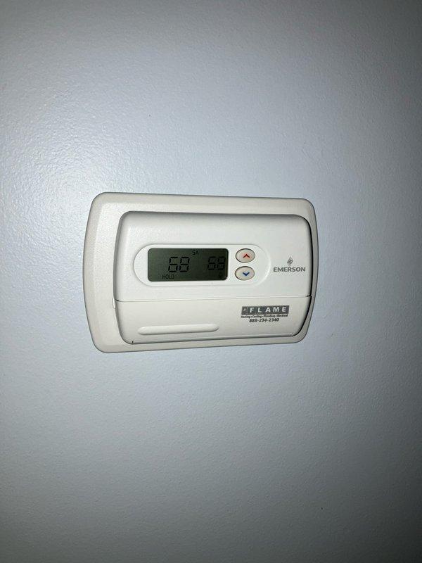 Completed assessment of non-operational A/C system over 20 years old. Observed significant weathering and oxidation on exterior electrical junction box with multiple conduits extending into ground cover. Interior inspection revealed Emerson digital thermostat (current reading 68°) mounted on interior wall with flame indicator visible. Customer seeking complete replacement of aging system.