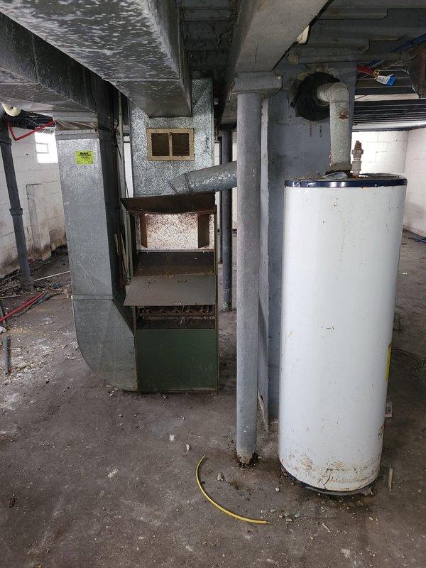 Conducted a full system estimate for furnace replacement in a residential basement. Inspection revealed an aging HVAC system with a green cabinet furnace showing significant wear alongside a cylindrical water heater with visible corrosion at its base. Observed deteriorating ductwork, moisture damage on ceiling surfaces, and recommended complete system replacement due to age and condition of equipment.