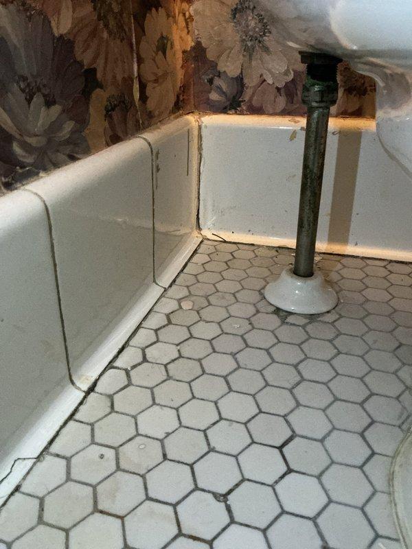 Completed inspection and repair of exposed plumbing infrastructure in bathroom area, including vertical PVC pipe installation with proper fittings and ventilation system connection. Addressed corner floor-to-wall junction where metal supply line emerges from hexagonal tile flooring, ensuring proper sealing and alignment with existing plumbing components.