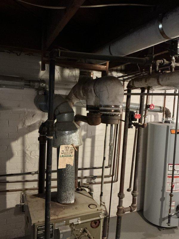 Conducted diagnostic assessment of leaking steam boiler system (likely original to property) located in basement. Boiler is currently operational despite leak. Inspected thermostat functionality and examined existing plumbing configuration including connected ductwork, flue pipe, and valve assemblies. Provided comprehensive replacement estimate for aging unit.
