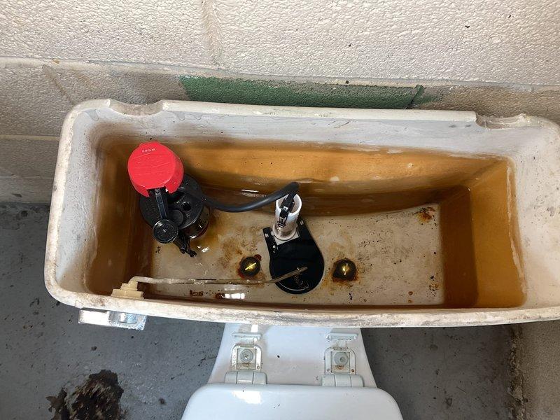 Responded to service call for a leaky water supply line at commercial toilet installation. Upon inspection, found corroded copper shut-off valve with visible water damage to surrounding concrete and significant mineral/rust buildup in tank with discolored water. Replaced damaged supply line and shut-off valve to resolve leak, then flushed system to remove sediment and restore proper function.