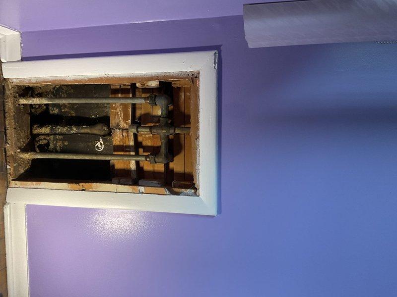Replaced corroded shower handle, shower head, and drain trap in bathroom with purple walls. Accessed plumbing through wall panel, revealing significant water damage and corrosion on copper supply lines. Completed installation of new components and verified proper function with no leaks.