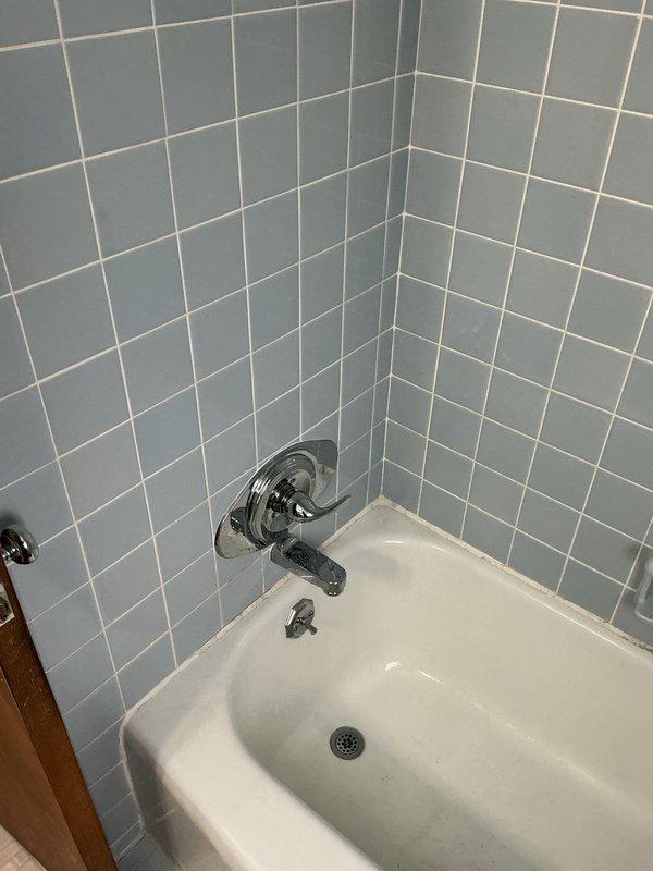 Accessed plumbing system through wall access panel to inspect copper supply line and mounting bracket. Performed maintenance on bathtub/shower fixture with blue tile surround, ensuring proper function of chrome faucet, controls, and drain.
