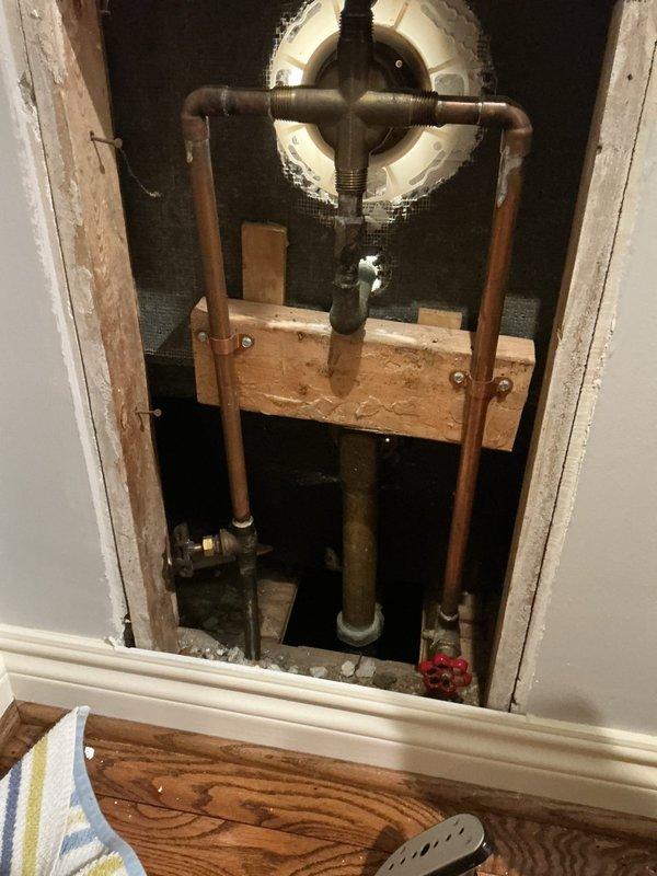 Performed inspection and assessment of shower fixture installation requirements in first floor bathroom of a 75-year-old residence. Examined existing plumbing infrastructure via access panel, revealing aged copper supply lines and supporting framework. Evaluated bathtub area which showed partially installed shower valve with visible water damage around plumbing openings in tile wall. Documentation completed with detailed photographs for comprehensive project planning.