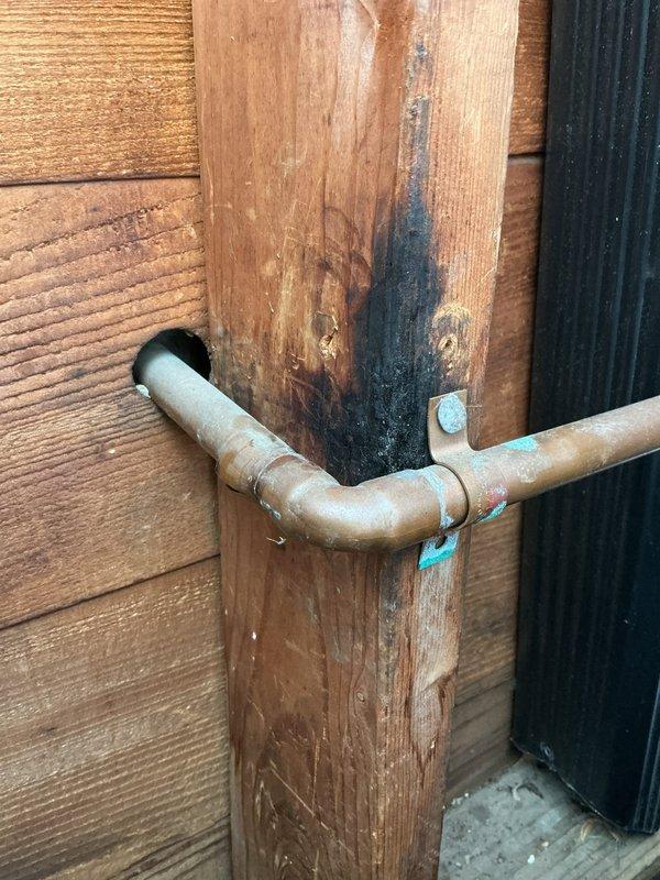 # Service Report: Water Leak Repair

Responded to service call for leaking outdoor spigot in garage with ruptured supply pipe causing water spray during operation. Upon inspection, identified severely corroded copper supply line with compromised elbow joint showing advanced oxidation and deterioration. The failed connection point was actively leaking when pressurized, with evidence of long-term moisture damage visible on adjacent wooden structural support, indicated by dark vertical staining and discoloration.

Replaced defective copper pipe section and corroded spigot assembly. Installed new frost-proof sillcock with proper mounting hardware on exterior wall. Verified all connections were secure, tested system under full pressure, and confirmed leak elimination with no further water discharge.