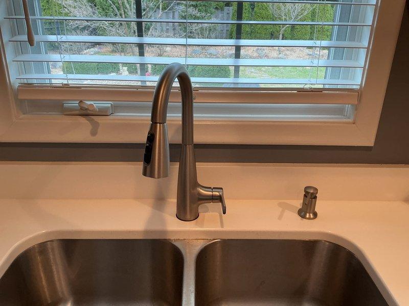 Completed installation of customer-supplied kitchen faucet and garbage disposal unit. Installed brushed nickel pull-down faucet with single lever handle above double-basin stainless steel undermount sink. Connected hot and cold water supply lines, ensuring proper water pressure and testing for leaks. Mounted matching soap dispenser on countertop deck. Installed garbage disposal unit beneath sink basin with proper electrical connection to air switch mounted on counter surface. Verified all components functioning properly with no leaks detected. System tested and operational upon completion.