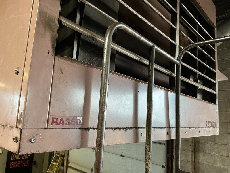Performed service call on Reznor RA350 oil-fired furnace unit located in commercial facility requiring ladder access. Inspected ceiling-mounted unit with horizontal louver discharge grille and verified proper safety railing installation around equipment. Documented unit condition and configuration, noting this is one of five total units on-site requiring future preventive maintenance scheduling. Unit accessible from ground level with ladder, positioned near corrugated metal ceiling structure with white ductwork distribution system.