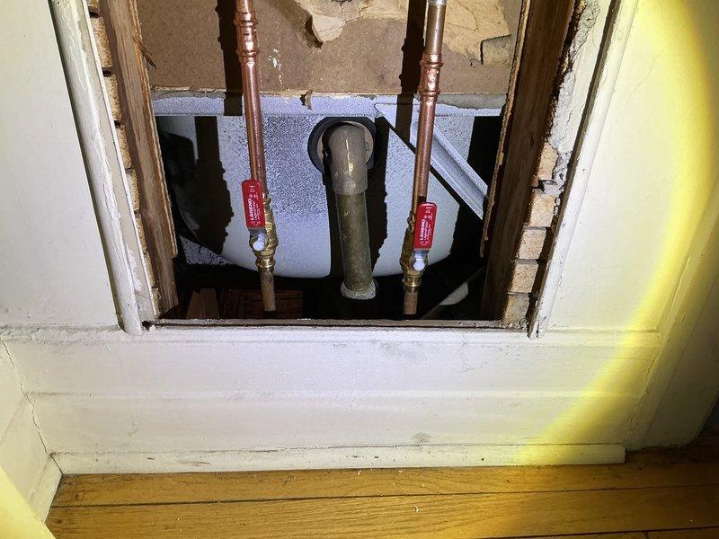 Responded to service call for water leaking from pipe in kitchen ceiling. Upon arrival, accessed plumbing infrastructure through existing wall opening at floor level. Identified source of leak in vertical copper supply line system serving kitchen fixtures above. Inspected two copper supply pipes with existing shut-off valves and central drain stack running through wall cavity. Evaluated valve integrity, pipe connections, and surrounding plumbing components for damage or deterioration. Created access point through drywall to expose affected plumbing for proper diagnosis and repair. Installed or verified operation of brass shut-off valves with red handles on both hot and cold water supply lines to isolate affected section. Addressed leak issue and restored water service to kitchen area. Documented conditions with photographs showing exposed copper supply lines, PVC drain stack, wall framing, and installed shut-off valve assembly for homeowner records.