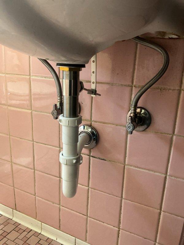 Completed installation of new chrome P-trap assembly and drain tailpiece under bathroom sink with pink ceramic tile surround. Replaced existing drainage components with white PVC P-trap configuration, ensuring proper alignment and secure connections to prevent leaks. Verified all chrome-plated fittings, braided supply lines, and wall-mounted shut-off valves were functioning correctly and properly secured. Tested drainage flow and checked all connections for leaks to confirm successful repair completion.