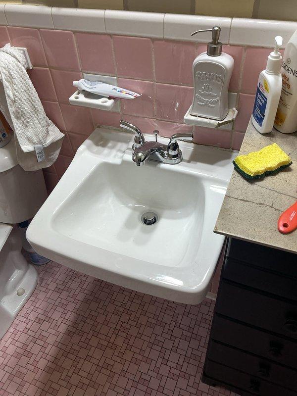 Completed installation of new chrome P-trap assembly and drain tailpiece under bathroom sink with pink ceramic tile surround. Replaced existing drainage components with white PVC P-trap configuration, ensuring proper alignment and secure connections to prevent leaks. Verified all chrome-plated fittings, braided supply lines, and wall-mounted shut-off valves were functioning correctly and properly secured. Tested drainage flow and checked all connections for leaks to confirm successful repair completion.