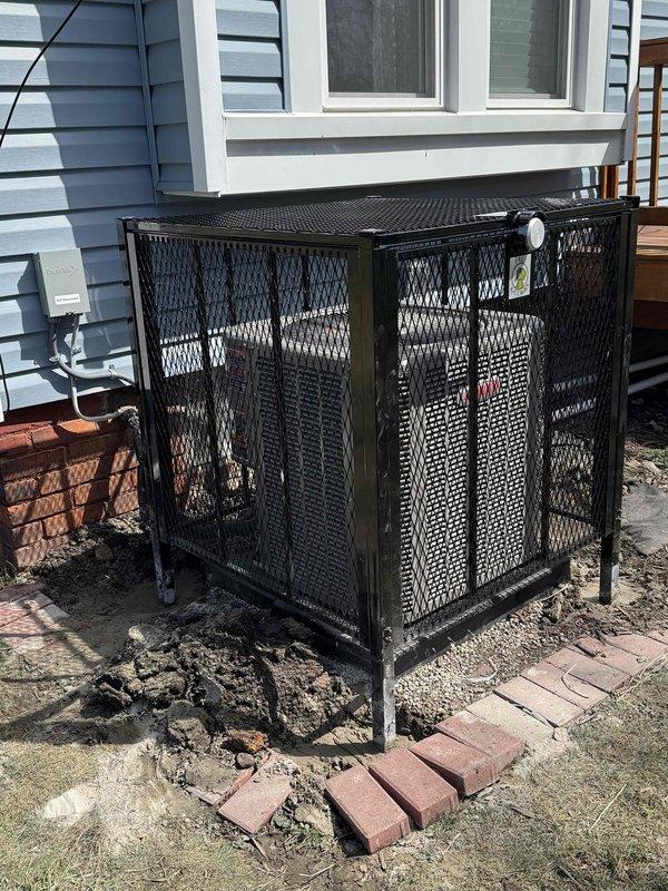 Installed black expanded metal mesh security cage enclosure for outdoor HVAC air conditioning unit on concrete pad adjacent to residential foundation. Cage features rectangular configuration with perforated mesh panels, flat top design, and access door panel to allow equipment servicing while providing protection and airflow. Unit positioned on metal legs next to blue-gray sided house with proper clearances maintained from foundation and glass block windows for adequate ventilation and code compliance.
