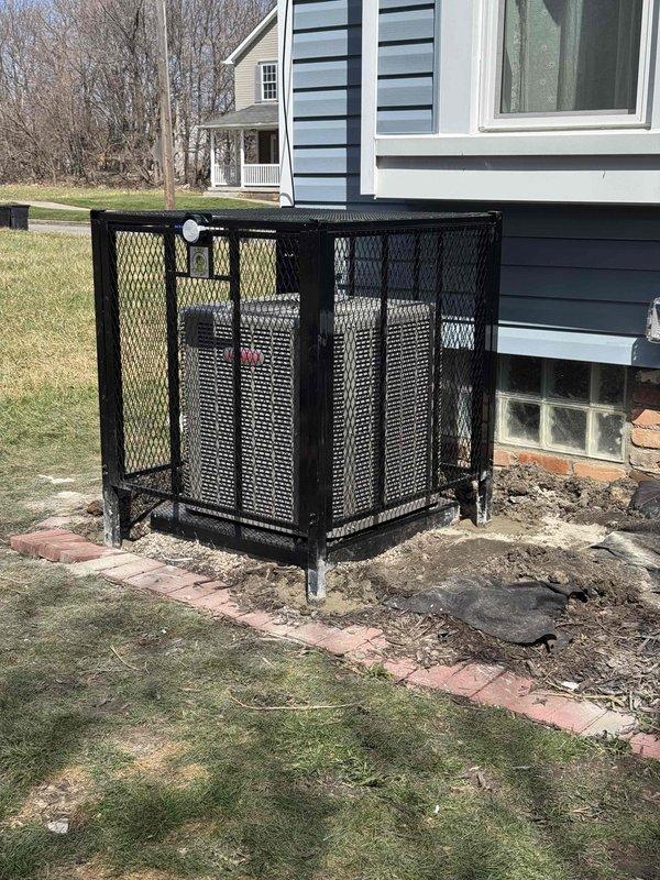 Installed black expanded metal mesh security cage enclosure for outdoor HVAC air conditioning unit on concrete pad adjacent to residential foundation. Cage features rectangular configuration with perforated mesh panels, flat top design, and access door panel to allow equipment servicing while providing protection and airflow. Unit positioned on metal legs next to blue-gray sided house with proper clearances maintained from foundation and glass block windows for adequate ventilation and code compliance.