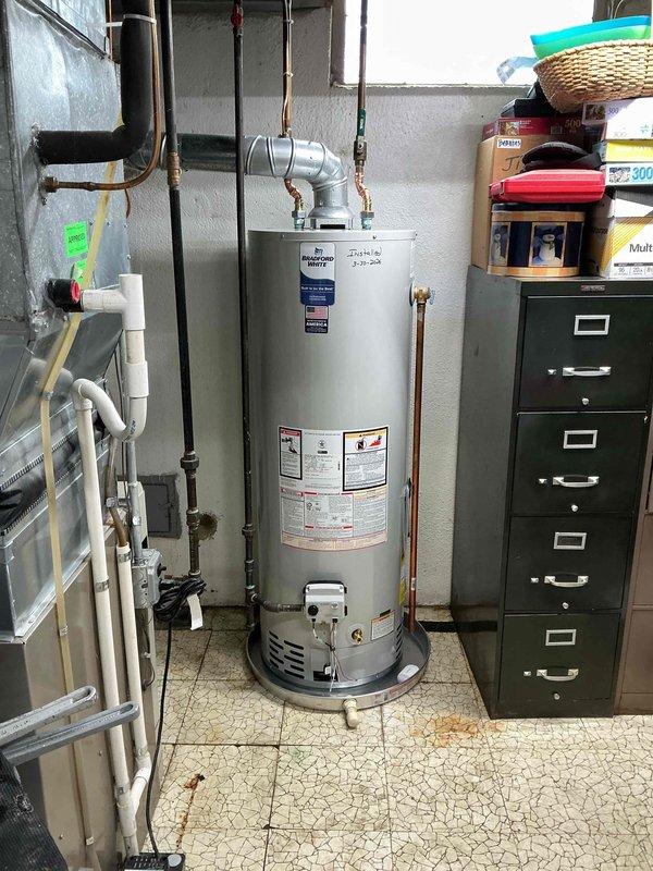 Installed a 40-gallon Bradford White natural draft atmospheric vent water heater (model RG240T6N) in basement utility area. Connected natural gas supply line, installed copper water piping connections, and configured proper venting system with dark metal vent pipes. Added required drain pan beneath unit to protect against potential water damage and ensure code compliance.