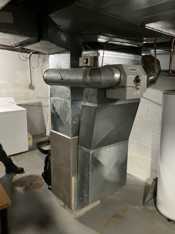 Completed furnace replacement estimate for basement installation with equipment age exceeding 10 years. Assessed existing gray metal cabinet furnace unit with horizontal ductwork and galvanized metal plenum connections. Documented current system configuration including ductwork transitions, utility room layout, and equipment accessibility for replacement planning. Photographed installation area showing concrete block walls, concrete flooring, and proximity to water heater to determine clearance requirements and proper equipment sizing for future replacement.