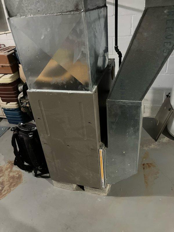 Completed furnace replacement estimate for basement installation with equipment age exceeding 10 years. Assessed existing gray metal cabinet furnace unit with horizontal ductwork and galvanized metal plenum connections. Documented current system configuration including ductwork transitions, utility room layout, and equipment accessibility for replacement planning. Photographed installation area showing concrete block walls, concrete flooring, and proximity to water heater to determine clearance requirements and proper equipment sizing for future replacement.