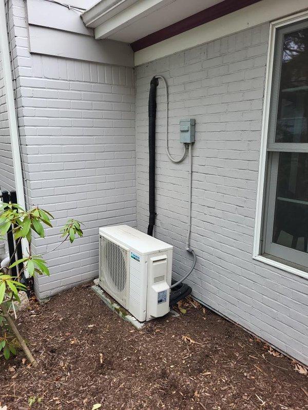 Evaluated existing 20-year-old Daikin mini split system with failed leaking coil requiring full system replacement. Assessed installation site for first-floor 3-season room application with outdoor condensing unit positioned on ground pad approximately 6 feet from indoor head location. Documented existing refrigerant line routing, electrical disconnect configuration, and exterior mounting conditions on white painted brick siding. Provided quote specifications for premium Daikin Atmosphera single-zone mini split system to replace failed equipment.