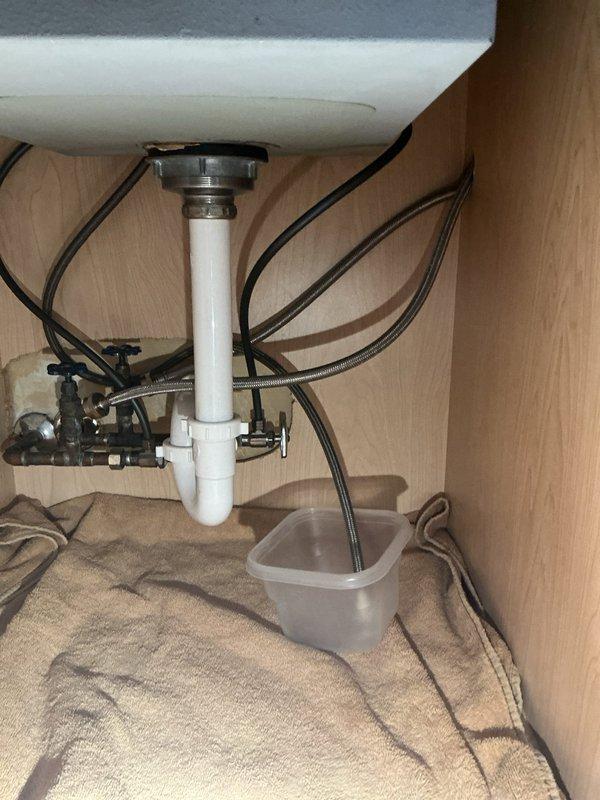 Responded to water leak complaint originating from laundry sink area. Upon arrival, conducted thorough inspection of sink plumbing system and cabinet interior. Examined P-trap assembly, drain connections, and supply line configuration for signs of active leakage or moisture damage. Verified integrity of chrome drain assembly, white PVC P-trap, and all connection points. Inspected black flexible supply hoses and shut-off valves for potential leak sources. Placed collection container beneath plumbing assembly as precautionary measure. Assessed undermount sink installation and faucet connections for any contributing factors. All plumbing components were evaluated and leak source was identified and addressed to restore proper function.