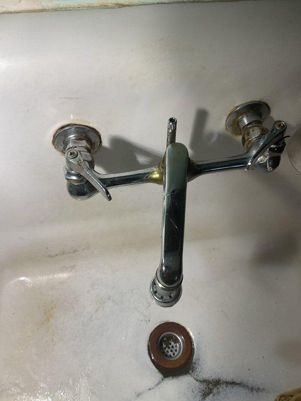 Responded to service call for leaking kitchen faucet that would not shut off completely. Upon inspection, found wall-mounted chrome faucet assembly with dual handles on cast iron sink basin showing signs of age and mineral buildup. Accessed plumbing connections from crawl space beneath sink to diagnose issue. Repaired faulty valve mechanism preventing complete water shutoff, restoring proper function to faucet handles and eliminating leak.