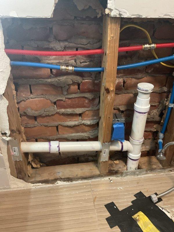 Responded to persistent sewage odor in bathroom of 1947-built home that began following previous drywall opening for leak repair behind toilet and washer/dryer area. Investigated exposed wall cavity revealing brick masonry construction with existing plumbing infrastructure including PVC drain/vent lines with visible purple primer at joints, flexible supply lines with brass fittings, and under-sink P-trap configuration with adjacent water filtration system. Inspected all visible plumbing connections, drain lines, and vent stack penetrations within the opened wall section to identify potential sewer gas escape points. Evaluated PVC joint integrity, trap seal functionality, and proper venting to determine source of eight-month odor issue confined to bathroom space.