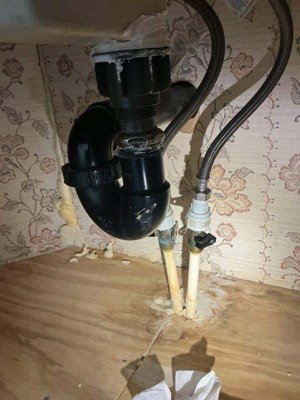 Responded to water leak under bathroom sink with reported mold odor in residence. Upon arrival, water supply had already been shut off for two weeks by homeowner. Performed diagnostic inspection of under-sink plumbing assembly, which consisted of black PVC P-trap configuration with flexible braided supply lines connected to aged white PVC supply pipes emerging from floor. Identified water damage and moisture staining on hardwood flooring surrounding pipe penetrations, along with mineral deposits and residue buildup at pipe bases. Observed deteriorated wallpaper and debris accumulation in affected area. Documented existing conditions including compromised shut-off valves with brass compression fittings and signs of prolonged moisture exposure on surrounding surfaces.