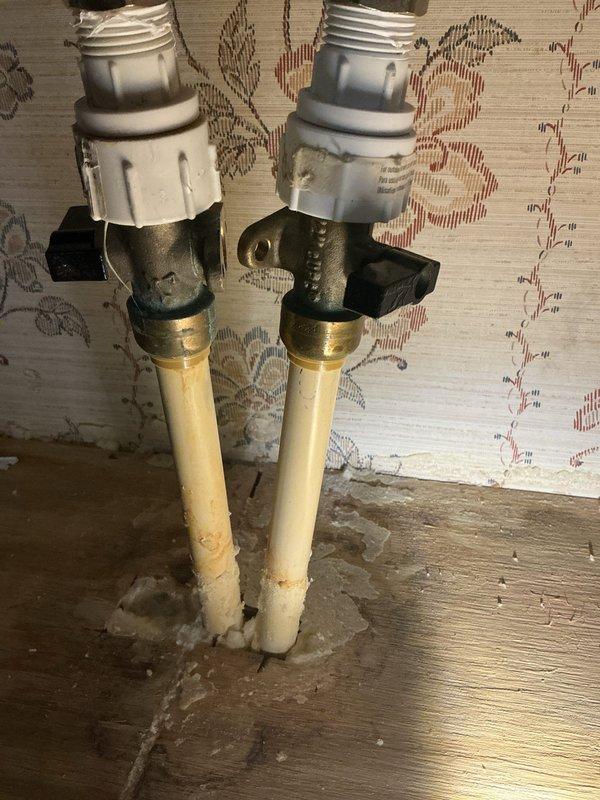 Responded to water leak under bathroom sink with reported mold odor in residence. Upon arrival, water supply had already been shut off for two weeks by homeowner. Performed diagnostic inspection of under-sink plumbing assembly, which consisted of black PVC P-trap configuration with flexible braided supply lines connected to aged white PVC supply pipes emerging from floor. Identified water damage and moisture staining on hardwood flooring surrounding pipe penetrations, along with mineral deposits and residue buildup at pipe bases. Observed deteriorated wallpaper and debris accumulation in affected area. Documented existing conditions including compromised shut-off valves with brass compression fittings and signs of prolonged moisture exposure on surrounding surfaces.