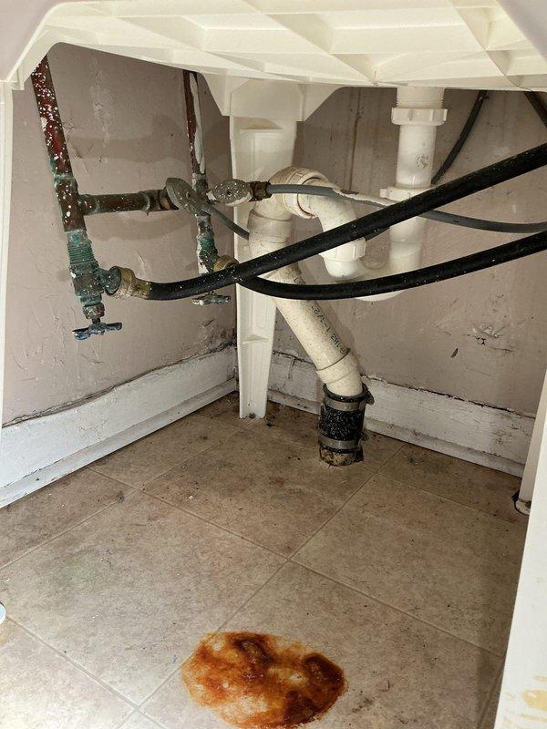 Responded to a service call for a corroded shut-off valve beneath the kitchen sink. Upon inspection, found heavily deteriorated metal piping with extensive oxidation, rust buildup, and copper corrosion presenting as greenish-blue patina. Evidence of water leakage was visible on the cabinet floor as brownish staining. The existing metal supply lines showed significant age-related degradation requiring replacement. Assessed the under-sink plumbing configuration which included a mix of white PVC drain piping with P-trap assembly and compromised metal supply components. Evaluated the shut-off valve condition and surrounding pipe infrastructure to determine appropriate repair or replacement options for the homeowner.