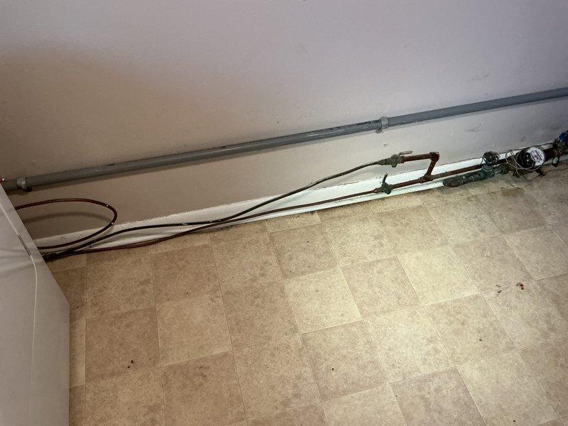 Responded to a service call for a corroded shut-off valve beneath the kitchen sink. Upon inspection, found heavily deteriorated metal piping with extensive oxidation, rust buildup, and copper corrosion presenting as greenish-blue patina. Evidence of water leakage was visible on the cabinet floor as brownish staining. The existing metal supply lines showed significant age-related degradation requiring replacement. Assessed the under-sink plumbing configuration which included a mix of white PVC drain piping with P-trap assembly and compromised metal supply components. Evaluated the shut-off valve condition and surrounding pipe infrastructure to determine appropriate repair or replacement options for the homeowner.