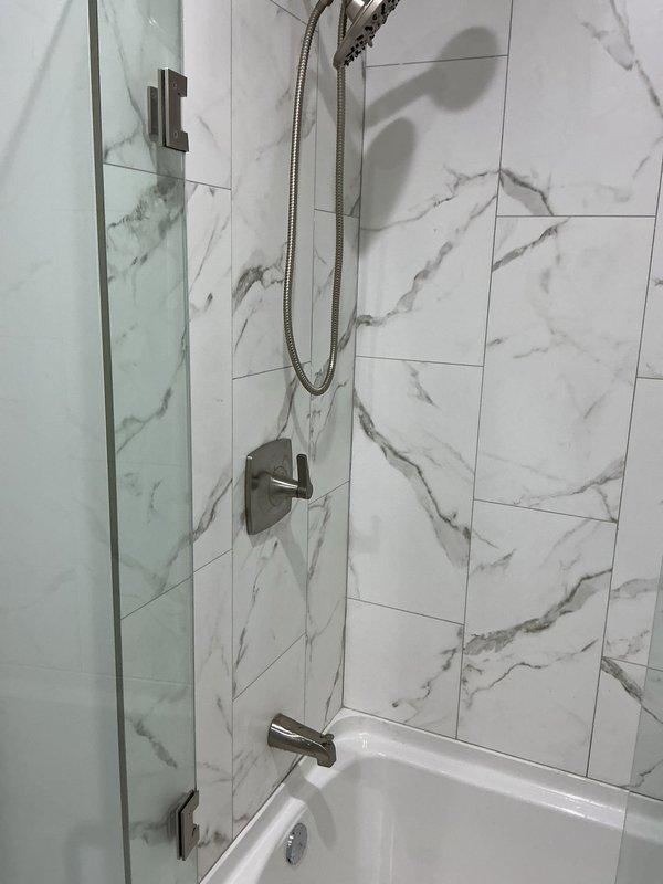 Completed standard showerhead installation in bathroom shower/tub combination unit. Installed handheld showerhead assembly with flexible braided metal hose and brushed nickel fixtures, coordinating with existing glass door hardware and tub spout. All plumbing connections were secured and tested for proper water flow and leak-free operation in the marble-patterned tile shower enclosure.