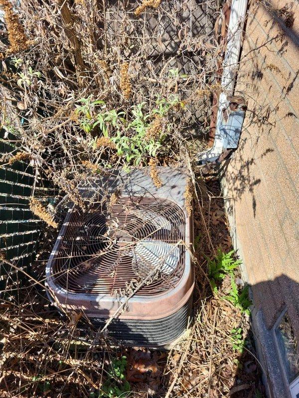 Conducted comprehensive site evaluation for full HVAC system replacement at residential property with significantly aged equipment. Existing outdoor condenser unit exhibited severe deterioration with extensive vegetation overgrowth, debris accumulation, and compromised airflow around unit perimeter. Equipment showed visible weathering, discoloration, and structural wear consistent with end-of-service-life conditions. Documented current system configuration including exterior condenser placement adjacent to building foundation with glass block basement windows and mixed brick-vinyl siding construction. Assessed clearance requirements and noted substantial vegetation management needed for proper equipment operation and future accessibility. Photographed existing installation conditions to support full system replacement estimate and equipment specifications.