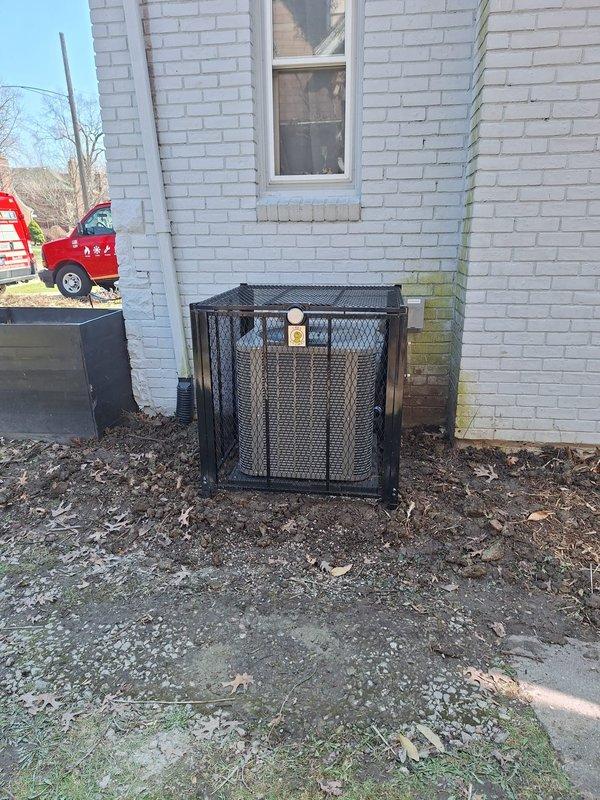 Installed protective black metal security cage around outdoor HVAC condenser unit at white brick building exterior. Cage features mesh screening on all sides with flat top cover and informational labeling, providing theft and vandalism protection while maintaining proper airflow for unit operation.