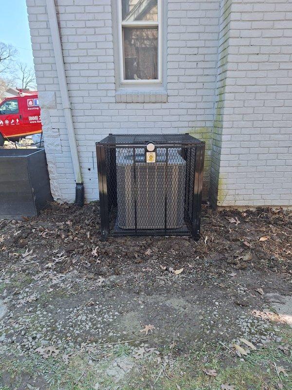 Installed protective black metal security cage around outdoor HVAC condenser unit at white brick building exterior. Cage features mesh screening on all sides with flat top cover and informational labeling, providing theft and vandalism protection while maintaining proper airflow for unit operation.