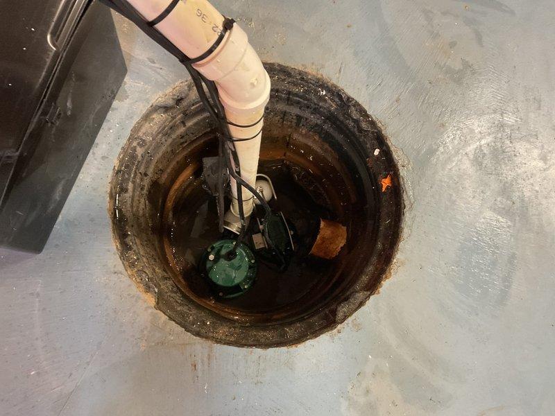 Replaced deteriorated sump pump system in basement pit showing extensive rust, mineral deposits, and corrosion from long-term water exposure. Installed new green submersible pump unit with float switch assembly, connected to existing white PVC discharge piping, and secured all electrical wiring and connections to restore proper water management functionality.