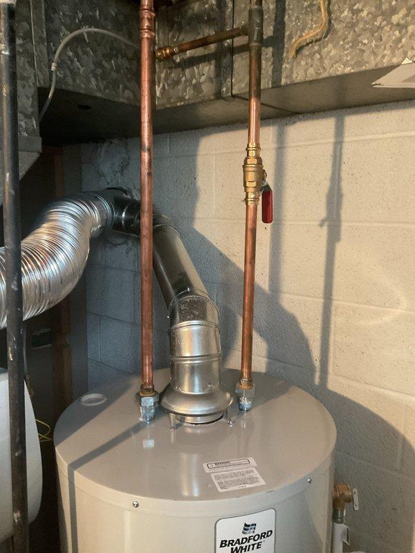 Completed installation of a 40-gallon short Bradford White tank water heater in basement utility area. Performed mechanical connections including copper supply piping with brass fittings, installed red-handled ball valve on hot water outlet, and mounted pressure relief valve on top of tank. Unit was positioned on checkered tile floor with white drain pan at base and reconnected all plumbing connections per manufacturer specifications and code requirements.