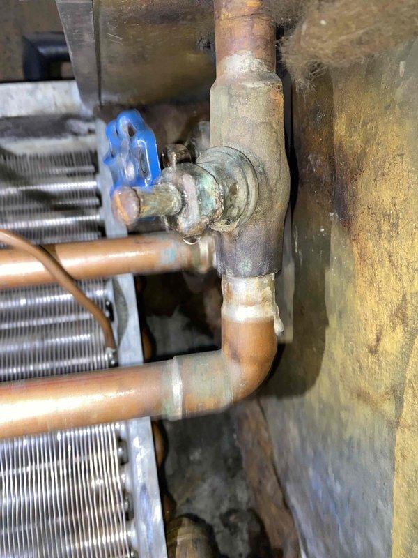 Responded to no heat call for 15-year-old boiler system in commercial unit. Upon inspection, identified leaking valve on copper piping assembly connected to heat exchanger coil, with significant corrosion and green patina indicating prolonged oxidation. Valve exhibited active leak with moisture accumulation in drain pan below unit. Documented existing conditions showing aged HVAC installation with deteriorated components and water damage to surrounding surfaces. Photographed corroded blue-handled valve, copper pipe connections, finned heat exchanger coil, and condensate management system for service records and repair recommendations.
