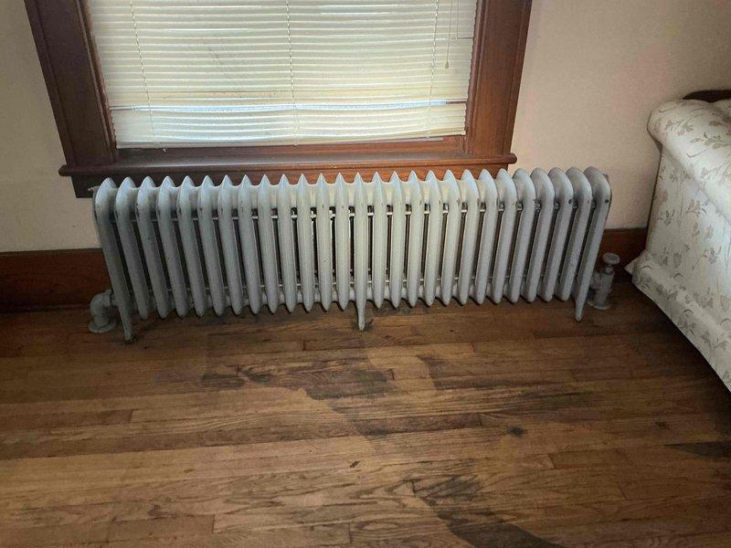 Responded to service call for burst radiator replacement on 3-year-old boiler system located in basement. Upon inspection, found white cast-iron radiator in living area with evidence of significant water damage to hardwood flooring beneath unit, indicating catastrophic failure. Assessed basement plumbing infrastructure and identified aged valve assembly with deteriorated paint and compromised fittings contributing to system failure. Documented extensive water staining on floor surfaces and deterioration of surrounding wooden structural components with exposed insulation. Performed diagnostic evaluation of radiator connections and associated piping to determine extent of damage and replacement requirements. Photographed affected radiator, water damage patterns, and basement valve assembly for documentation and repair planning purposes.