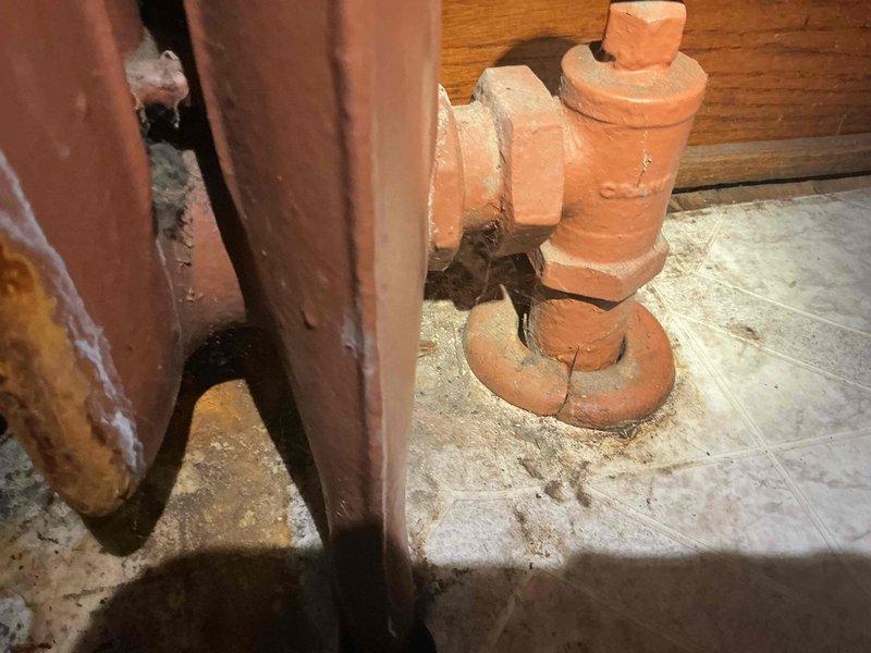 Responded to service call for burst radiator replacement on 3-year-old boiler system located in basement. Upon inspection, found white cast-iron radiator in living area with evidence of significant water damage to hardwood flooring beneath unit, indicating catastrophic failure. Assessed basement plumbing infrastructure and identified aged valve assembly with deteriorated paint and compromised fittings contributing to system failure. Documented extensive water staining on floor surfaces and deterioration of surrounding wooden structural components with exposed insulation. Performed diagnostic evaluation of radiator connections and associated piping to determine extent of damage and replacement requirements. Photographed affected radiator, water damage patterns, and basement valve assembly for documentation and repair planning purposes.